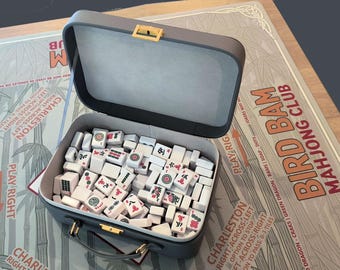 Op de afbeelding: Een open grijze Mahjong-koffer gevuld met witte tegels met rode en zwarte symbolen. De koffer staat op een spelbord met de tekst "Bird Bam Mahjong Club".