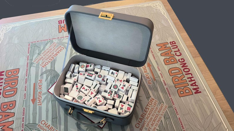 Puede incluir: Un estuche de Mahjong gris abierto, lleno de fichas blancas con s&iacute;mbolos rojos y negros. El estuche est&aacute; sobre un tablero de juego con el texto "Bird Bam Mahjong Club".