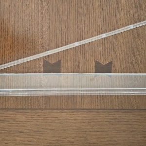 May include: A clear acrylic refrigerator door shelf bin featuring a long, rectangular storage compartment and an attached, angled support arm with a hook mechanism on the end for mounting inside a refrigerator door interior.
