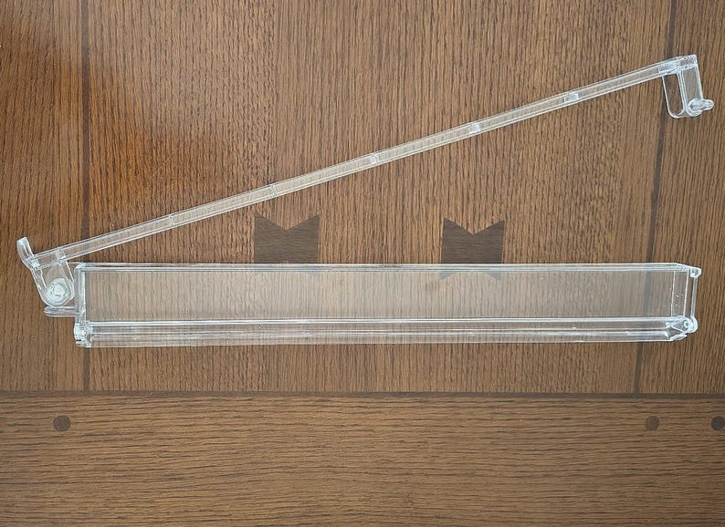 May include: A clear acrylic refrigerator door shelf bin featuring a long, rectangular storage compartment and an attached, angled support arm with a hook mechanism on the end for mounting inside a refrigerator door interior.