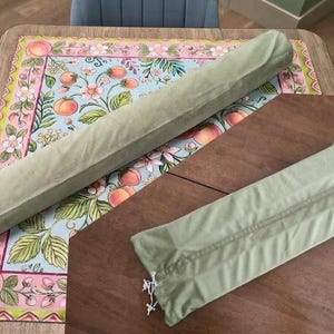 May include: Two sage green fabric storage sleeves with white drawstring closures. One sleeve is long and cylindrical, while the other is shorter and rectangular. Both items feature a soft, matte texture and are designed for organizing or protecting items.