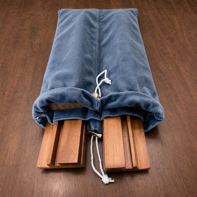 May include: A soft, slate blue velvet drawstring pouch containing two rectangular wooden blocks. The pouch features a white cord with a small metallic toggle, and the wooden blocks are visible at the opening, showing a smooth, natural finish.