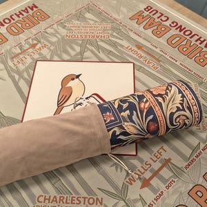 K&ouml;nnte beinhalten: Eine aufgerollte Mahjong-Matte mit floralem Design ist teilweise sichtbar, zusammen mit einer passenden Tragetasche. Eine Karte mit einer Vogelillustration liegt auf der Matte. Die Matte liegt auf einem Tisch mit dem Aufdruck "Bird Bam Mahjong Club".