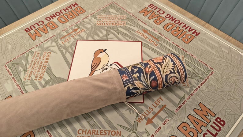 K&ouml;nnte beinhalten: Eine aufgerollte Mahjong-Matte mit floralem Design ist teilweise sichtbar, zusammen mit einer passenden Tragetasche. Eine Karte mit einer Vogelillustration liegt auf der Matte. Die Matte liegt auf einem Tisch mit dem Aufdruck "Bird Bam Mahjong Club".