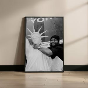 May include: Black and white framed print featuring a person holding a Statue of Liberty replica. The person is wearing a black beanie and a black shirt. The background has the word "York" visible.