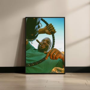 May include: Framed print featuring a smiling person holding a cigar and a steering wheel-like object. The person is wearing a green shirt. The background shows a blue sky and a glimpse of a landscape.
