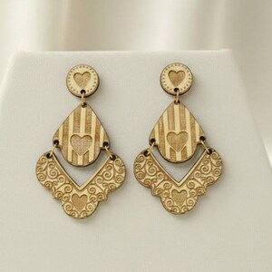 May include: A pair of wooden earrings with a heart and striped design. The earrings have a circular top, a teardrop-shaped middle section with vertical stripes, and a decorative bottom with a heart-shaped cutout. The earrings are light brown.