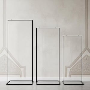 May include: Three black metal display stands of varying heights. Each stand has a rectangular base and a rectangular frame. The stands are empty and set against a neutral-toned wall with a subtle pattern.