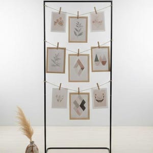 Könnte beinhalten: Ein schwarzer Metall-Fotoständer mit neun gerahmten Drucken. Die Drucke zeigen botanische und abstrakte Designs, die mit Holzklammern an Schnüren befestigt sind. Im Vordergrund steht eine Vase mit getrocknetem Pampasgras.