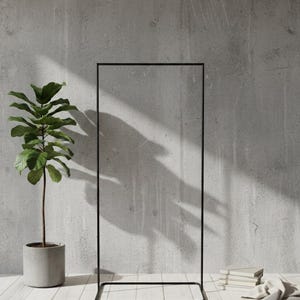 Può includere: Un appendiabiti in metallo nero contro un muro di cemento grigio. Una pianta in vaso con foglie verdi è a sinistra, e una pila di libri e un panno piegato sono a destra. L'appendiabiti ha una cornice rettangolare.