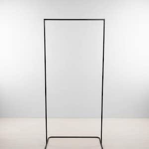 May include: A tall, black metal clothing rack with a rectangular base and a rectangular top. The rack is empty and stands against a white wall and light wood floor.