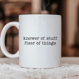 May include: A white ceramic mug with the text "knower of stuff fixer of things" in black. The mug is on a white, textured coaster, with steam rising from the top. The background is blurred.