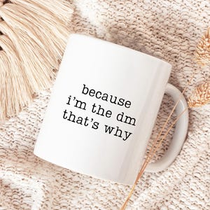 May include: White ceramic mug with the text "because i'm the dm that's why" in black. The mug is on a cream-colored knitted surface, with a beige macrame and dried wheat stalks in the background.