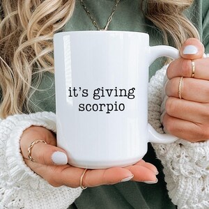 May include: White ceramic mug with the text "it's giving scorpio" in black. The mug is held in front of a person wearing a green shirt and a white sweater. The mug is a standard size.