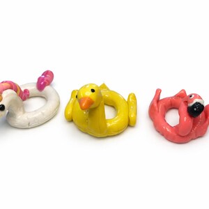 May include: Three miniature pool floaties: a white unicorn with a pink and orange mane, a yellow duck, and a pink flamingo with a black beak. These small, colorful floaties are likely for decorative purposes.