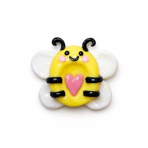 May include: A yellow bee-shaped decorative item with a pink heart, black stripes, and white wings. The bee has a smiling face with black antennae. The item is likely a brooch, pin, or decorative element.