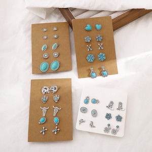 May include: Multiple sets of stud earrings on display. Each set features silver-toned metal and turquoise-colored stones in various shapes, including hearts, flowers, and crosses. Some sets have a western theme with cowboy boots and bull skulls.