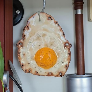 May include: A ceramic fried egg ornament with a bright yellow yolk and speckled seasoning. The egg white has a slightly browned, textured edge. It hangs from a metal hook, suggesting kitchen or food-themed decor.