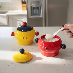 以下が含まれることがあります： 蓋とスプーンが付いた3つのセラミックボウルのセット。ボウルは黄色、赤、青で、赤と青のアクセントが付いています。黄色のボウルには青い蓋があり、赤いボウルには白い物質が入っています。