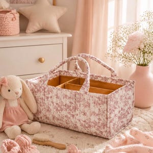 Peut inclure: Un panier de rangement rectangulaire en tissu matelassé avec un motif floral toile de Jouy rose poudré sur fond blanc. L'intérieur est doublé d'un tissu jaune moutarde uni et divisé en trois sections avec deux anses en tissu robustes.