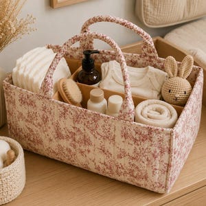 Peut inclure: Un panier de rangement rectangulaire en tissu avec un motif floral rouge sur fond crème et deux anses. L'intérieur est divisé en compartiments contenant des tissus blancs pliés, un flacon sombre, deux petits flacons blancs, une brosse et un lapin en crochet.