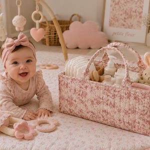 Peut inclure: Un panier de rangement rectangulaire en tissu matelassé avec un motif floral rose et blanc et deux anses robustes. Le panier contient des articles de puériculture, notamment des couches blanches, des petits flacons et un lapin en crochet beige.