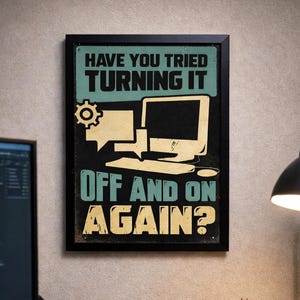 Puede incluir: Un escritorio con un monitor de ordenador, un teclado y un cartel enmarcado con el texto "¿Has probado a apagarlo y volverlo a encender?". También hay un teléfono, una taza y una lámpara de escritorio. Iluminación cálida.