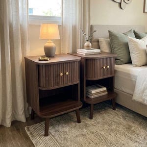 May include: Two dark brown wooden nightstands with a ribbed design on the drawer fronts and gold-colored knobs. Each nightstand has a lower shelf and is set on four legs. A lamp and decorative items sit on top of one.