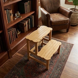 May include: A two-step wooden step stool with a scalloped edge design. The light-colored bamboo stool is placed on a patterned rug in front of a bookshelf and a leather armchair. The step stool is designed to help reach higher shelves.