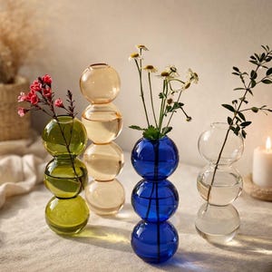 Peut inclure: Plusieurs vases décoratifs en verre de différentes couleurs, dont vert, ambre, bleu et transparent. Chaque vase est composé de sections sphériques empilées et contient des fleurs fraîches et de la verdure.