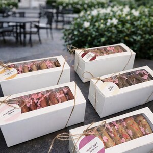 K&ouml;nnte beinhalten: Mehrere wei&szlig;e Schachteln mit transparenten Fenstern, jede mit Bindfaden gebunden und mit Keksen gef&uuml;llt. Die Schachteln sind mit Geschmacksrichtungen wie "Strawberry Creme" und "Classic Sugar Sprinkle" beschriftet. Rosa Papierschnipsel sind in den Schachteln sichtbar.
