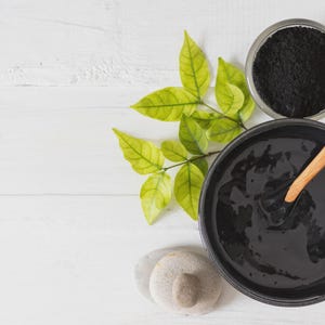 Puede incluir: Vista superior de una composición para el cuidado de la piel con una mascarilla negra en un cuenco y una cuchara de madera. También hay un vaso pequeño con polvo negro y una ramita de hojas verdes. También se incluyen dos piedras pequeñas y lisas.