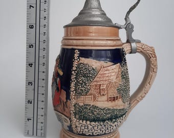 May include: A ceramic beer stein with a hinged metal lid. The stein features a detailed relief scene of a village with figures and buildings. The handle is intricately designed. The stein is approximately 9 inches tall.