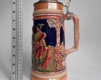 May include: A ceramic beer stein with a detailed hunting scene in relief. The stein features a hinged metal lid, a decorative handle, and the word "GUTE" on the side. The colors include brown, blue, and cream.