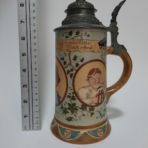 May include: A ceramic beer stein with a metal lid and handle, featuring illustrations of people and the German text 'Ein guter kühler Trunk erfreut!'. A ruler placed next to the stein indicates a height of approximately 9.5 inches.