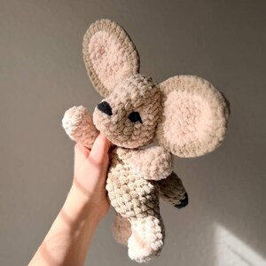 以下が含まれることがあります： ベージュと茶色の手作りのぬいぐるみマウスのおもちゃ。 マウスは、大きくて丸い耳、小さな黒い鼻、足に黒いアクセントがあります。 柔らかく、質感のある素材が、おもちゃに抱きしめたくなるような外観を与えています。