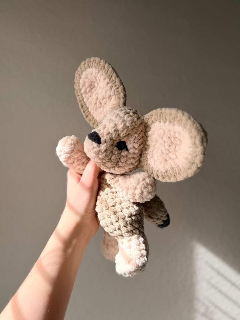 以下が含まれることがあります： ベージュと茶色の手作りのぬいぐるみマウスのおもちゃ。 マウスは、大きくて丸い耳、小さな黒い鼻、足に黒いアクセントがあります。 柔らかく、質感のある素材が、おもちゃに抱きしめたくなるような外観を与えています。