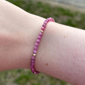 May include: A delicate bracelet featuring small, round, pink gemstones strung together. The bracelet is accented with small gold-colored beads. The jewelry is worn on a wrist, showcasing the bracelet's design and color.