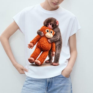 Puede incluir: Camiseta blanca con un gráfico de un mono marrón abrazando a un mono de peluche naranja. El juguete de peluche tiene una cara sonriente. La camiseta la lleva una persona con vaqueros azules.