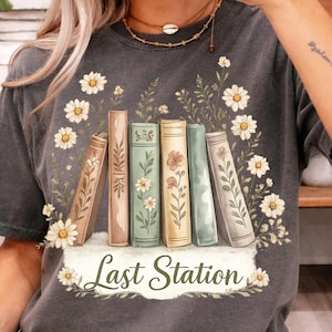 May include: Dark gray t-shirt featuring a floral design with daisies and greenery. The graphic includes a row of vintage books with floral accents and the text "Last Station" in a decorative font.