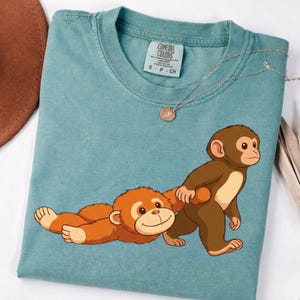 Puede incluir: Una camiseta Comfort Colors de color verde azulado con una ilustración de dibujos animados de dos monos. Un mono está acostado y el otro caminando. Un collar plateado con un colgante está sobre la camisa.