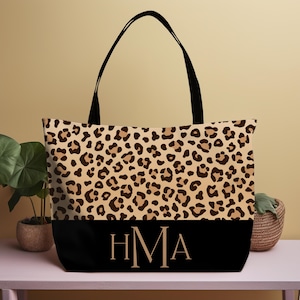 May include: A large tote bag with a leopard print pattern on the upper portion and a solid black bottom. The bag has black handles and the initials "HMA" in gold lettering on the black section. The bag is sitting on a pink surface.