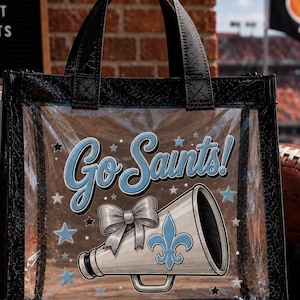 May include: A clear plastic tote bag with black trim and black handles. The front features the text Go Saints! in light blue, a white megaphone with a light blue fleur-de-lis, a silver bow, and small light blue and silver stars printed on the surface.