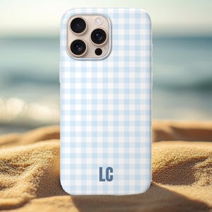 May include: A light blue and white gingham pattern phone case with the initials "LC" in the lower center. The case has a glossy finish and is shown on a sandy surface with a blurred ocean background.
