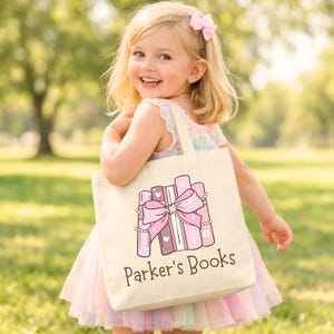 May include: A canvas tote bag with a pink bow and books design, with the text "Parker's Books". The bag is cream-colored and has a rectangular shape. The design features pink and purple books with a large pink bow.