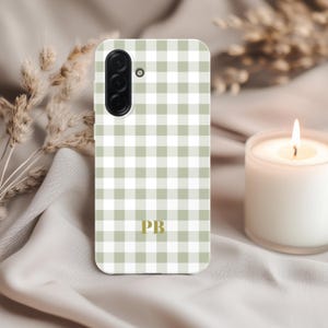 May include: A phone case with a green and white gingham pattern. The case has a camera and flash cutout at the top and gold-colored initials "PB" at the bottom. The case is on a light-colored surface with a lit candle and dried plants in the background.