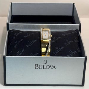 May include: A gold-tone Bulova wristwatch with a rectangular face and a crystal-accented bezel and band. The watch is presented on a black branded pillow inside a silver and black Bulova box with the brand name and logo printed on the front.