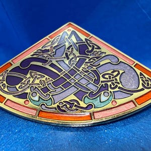May include: A fan-shaped metal brooch featuring intricate gold-toned linework filled with enamel in shades of deep purple, lavender, teal, and orange. The design displays a complex, interwoven knotwork pattern with symmetrical, stylized animal motifs.