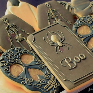 May include: Assortment of Halloween-themed cookies. The cookies feature designs such as skeletons, sugar skulls, bats, and a rectangle cookie with a spider and the word "Boo". The cookies are decorated with dark gray, gold, and orange icing.