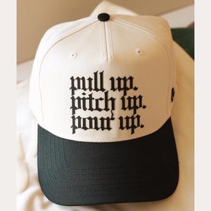 May include: A white baseball cap with a black brim. The cap features the text "pull up. pitch up. pour up." in a gothic font. The cap has a black button on top.